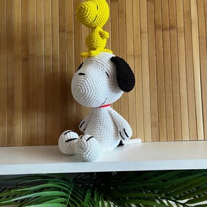Pode incluir: Um brinquedo de pel&uacute;cia Snoopy e Woodstock feito &agrave; m&atilde;o em croch&ecirc;. Snoopy &eacute; branco com orelhas pretas e uma coleira vermelha, enquanto Woodstock &eacute; amarelo. As figuras est&atilde;o sentadas em uma superf&iacute;cie branca contra um fundo de madeira. Os brinquedos de pel&uacute;cia s&atilde;o detalhados com detalhes costurados.