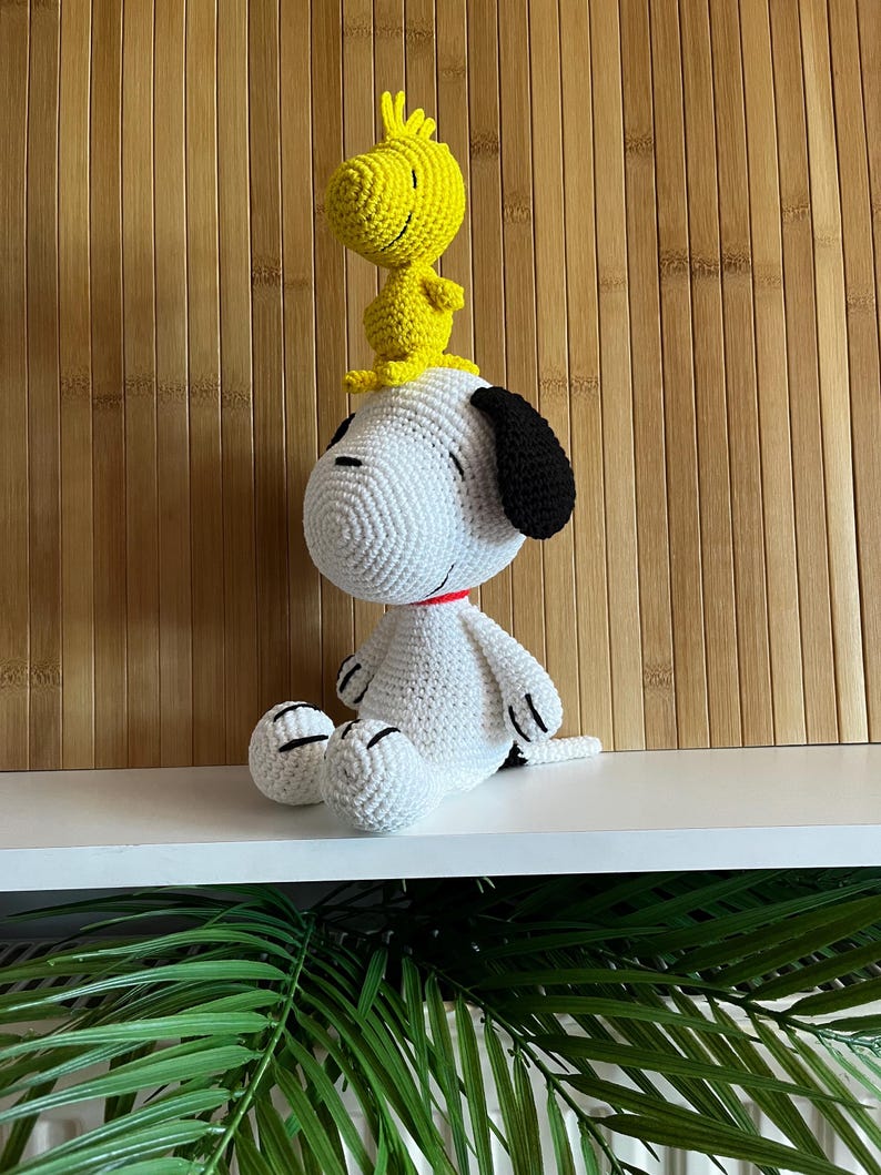 Pode incluir: Um brinquedo de pel&uacute;cia Snoopy e Woodstock feito &agrave; m&atilde;o em croch&ecirc;. Snoopy &eacute; branco com orelhas pretas e uma coleira vermelha, enquanto Woodstock &eacute; amarelo. As figuras est&atilde;o sentadas em uma superf&iacute;cie branca contra um fundo de madeira. Os brinquedos de pel&uacute;cia s&atilde;o detalhados com detalhes costurados.