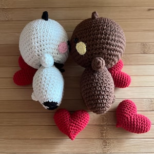 Könnte beinhalten: Gehäkelte Figuren von zwei Charakteren, einer weißen und einer braunen, mit roten Flügeln und Herzen. Die weiße Figur hat einen schwarzen Haarknoten und rosa Wangen. Die braune Figur hat eine gelbe Wange. Drei rote gehäkelte Herzen sind ebenfalls vorhanden.