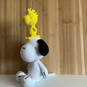 Pode incluir: Um brinquedo de pel&uacute;cia Snoopy e Woodstock feito &agrave; m&atilde;o em croch&ecirc;. Snoopy &eacute; branco com orelhas pretas e Woodstock &eacute; amarelo. Snoopy est&aacute; sentado com Woodstock empoleirado em sua cabe&ccedil;a. Os brinquedos t&ecirc;m detalhes em vermelho e preto.