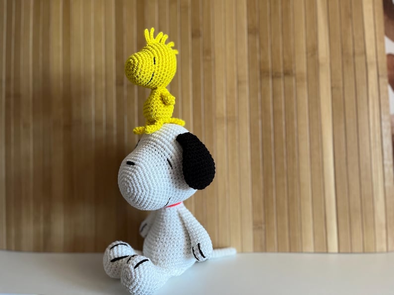 Pode incluir: Um brinquedo de pel&uacute;cia Snoopy e Woodstock feito &agrave; m&atilde;o em croch&ecirc;. Snoopy &eacute; branco com orelhas pretas e Woodstock &eacute; amarelo. Snoopy est&aacute; sentado com Woodstock empoleirado em sua cabe&ccedil;a. Os brinquedos t&ecirc;m detalhes em vermelho e preto.