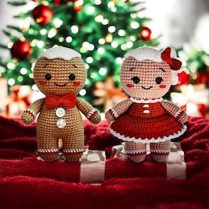 Op de afbeelding: Twee handgemaakte gehaakte peperkoeken poppen. Eén is een peperkoekenman met een rode strik en knopen. De andere is een peperkoekenvrouw met een rode jurk met witte rand en een strik. Ze staan op een rode ondergrond met een kerstboom op de achtergrond.