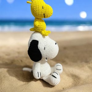 Pode incluir: Figuras de croch&ecirc; de Snoopy e Woodstock sentadas em uma praia arenosa com o oceano e o c&eacute;u azul ao fundo. Snoopy &eacute; branco com orelhas pretas, e Woodstock &eacute; amarelo. As figuras s&atilde;o feitas &agrave; m&atilde;o.