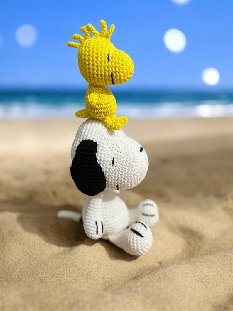 Pode incluir: Figuras de croch&ecirc; de Snoopy e Woodstock sentadas em uma praia arenosa com o oceano e o c&eacute;u azul ao fundo. Snoopy &eacute; branco com orelhas pretas, e Woodstock &eacute; amarelo. As figuras s&atilde;o feitas &agrave; m&atilde;o.