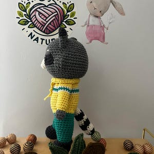 May include: A crocheted raccoon toy wearing a yellow shirt with green stripes and green pants. The toy has a black and white striped tail. The toy is standing on a wooden surface with green leaves and colorful balls.