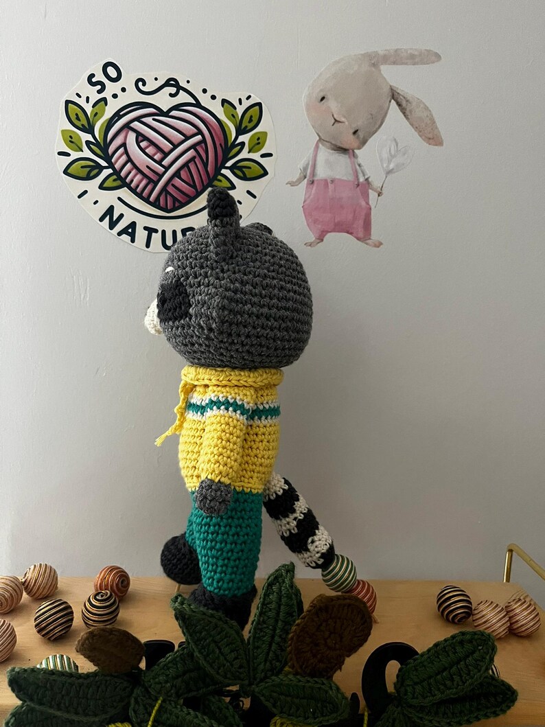 May include: A crocheted raccoon toy wearing a yellow shirt with green stripes and green pants. The toy has a black and white striped tail. The toy is standing on a wooden surface with green leaves and colorful balls.