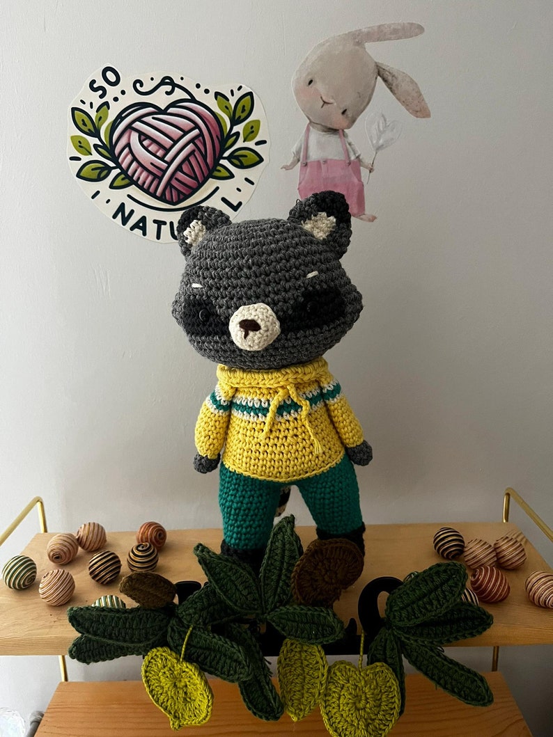 May include: A crocheted raccoon wearing a yellow and green sweater and green pants. The raccoon is standing on a wooden shelf with a green leafy vine and brown and gold striped balls.