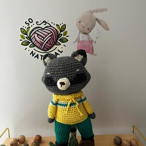 May include: A crocheted raccoon plush toy wearing a yellow and green sweater and pants. The toy is standing on a wooden surface with green leaves and brown and white striped balls.
