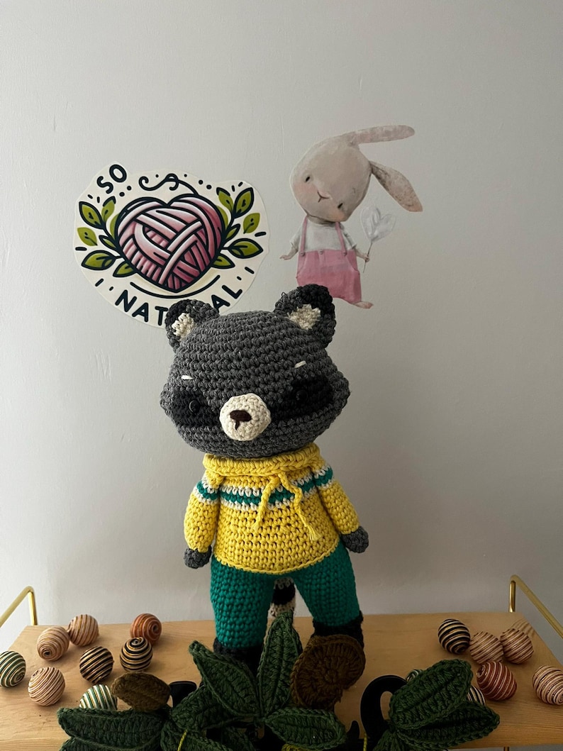 May include: A crocheted raccoon plush toy wearing a yellow and green sweater and pants. The toy is standing on a wooden surface with green leaves and brown and white striped balls.