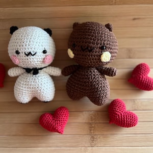 Może przedstawiać: Dwie szydełkowe figurki amigurumi: biała panda z czarnymi akcentami i brązowy miś, każdy z różowymi policzkami. Czerwone szydełkowe serca są rozrzucone wokół nich. Figurki znajdują się na jasnobrązowej drewnianej powierzchni.