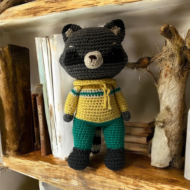 May include: A crocheted raccoon plush toy wearing a yellow and green sweater and green pants. The toy is sitting on a wooden shelf with books and a branch.