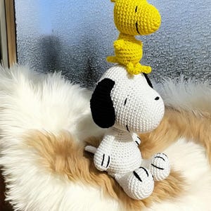 Pode incluir: Figuras de Snoopy e Woodstock em croch&ecirc;. Snoopy &eacute; branco com orelhas pretas e Woodstock &eacute; amarelo. Est&atilde;o sentados numa superf&iacute;cie fofa de cor creme.
