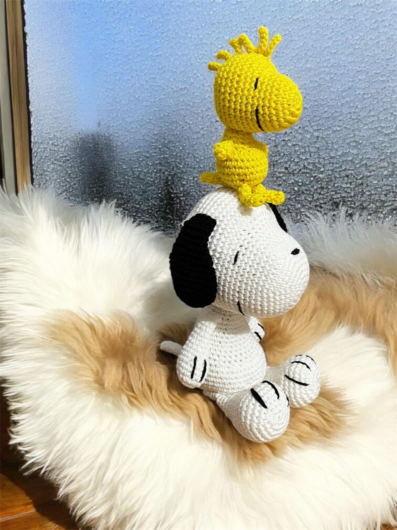 Pode incluir: Figuras de Snoopy e Woodstock em croch&ecirc;. Snoopy &eacute; branco com orelhas pretas e Woodstock &eacute; amarelo. Est&atilde;o sentados numa superf&iacute;cie fofa de cor creme.