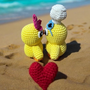 Puede incluir: Dos juguetes de pollitos amarillos tejidos a crochet con detalles en rosa y azul se besan en una playa de arena. Un corazón rojo tejido a crochet está en primer plano.