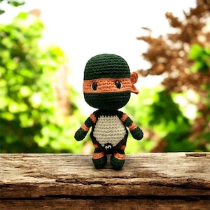 Puede incluir: Muñeca de ganchillo hecha a mano de un personaje de tortuga ninja. La muñeca tiene una cubierta para la cabeza verde con una máscara naranja y una raya naranja a juego en los brazos y las piernas. El cuerpo es de color canela con un contorno marrón. La muñeca está sobre una superficie de madera.