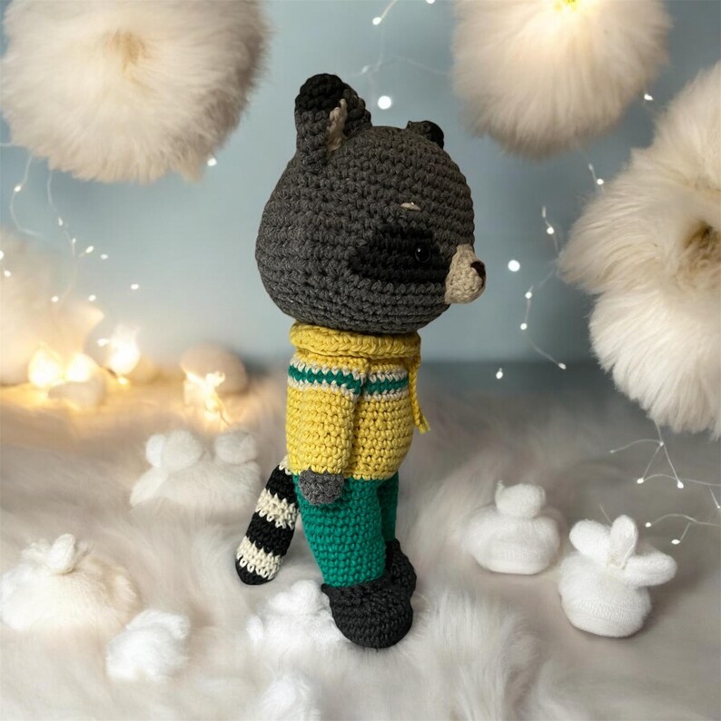 May include: A crocheted raccoon toy wearing a yellow sweater and green pants. The toy has a black and white striped tail and is standing on a white fluffy surface.
