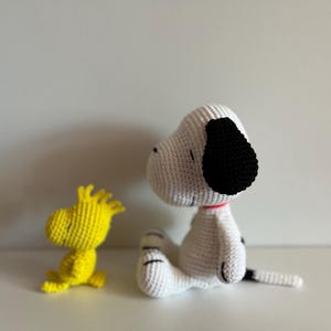 Pode incluir: Figuras de Snoopy e Woodstock de croch&ecirc;. Snoopy &eacute; branco com orelhas pretas e uma mancha preta nas costas, usando uma coleira vermelha. Woodstock &eacute; amarelo. Ambos est&atilde;o sentados em uma superf&iacute;cie branca.