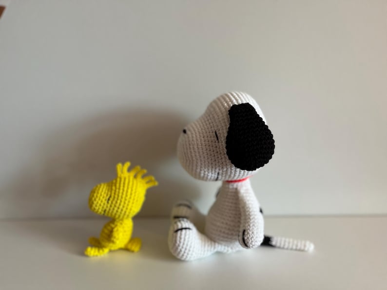 Pode incluir: Figuras de Snoopy e Woodstock de croch&ecirc;. Snoopy &eacute; branco com orelhas pretas e uma mancha preta nas costas, usando uma coleira vermelha. Woodstock &eacute; amarelo. Ambos est&atilde;o sentados em uma superf&iacute;cie branca.