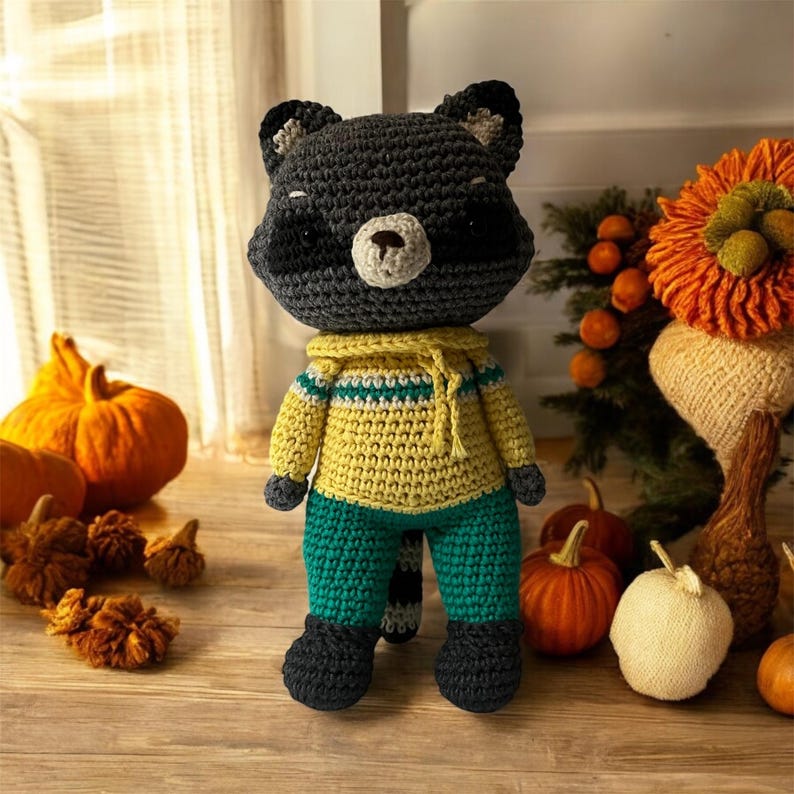 May include: A crocheted raccoon plush toy wearing a yellow and green sweater and pants. The toy is standing on a wooden surface with pumpkins and other fall decorations.