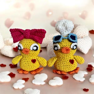 May include: Two handmade yellow crochet ducklings with orange beaks and feet. One duckling has a large pink bow, the other wears a chef's hat and blue goggles. Both have a red heart on their chest.