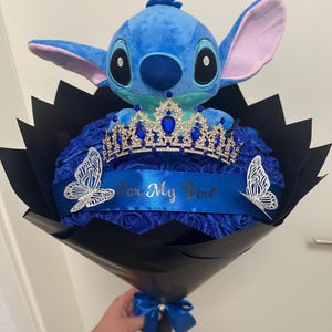 Könnte beinhalten: Ein blauer Stitch-Plüschtier mit einer Juwelenkrone auf einem Strauß blauer Rosen. Der Strauß ist in schwarzes Papier gewickelt und mit einem blauen Band und Schmetterlingen verziert. Ein blaues Banner liest "For My Girl."