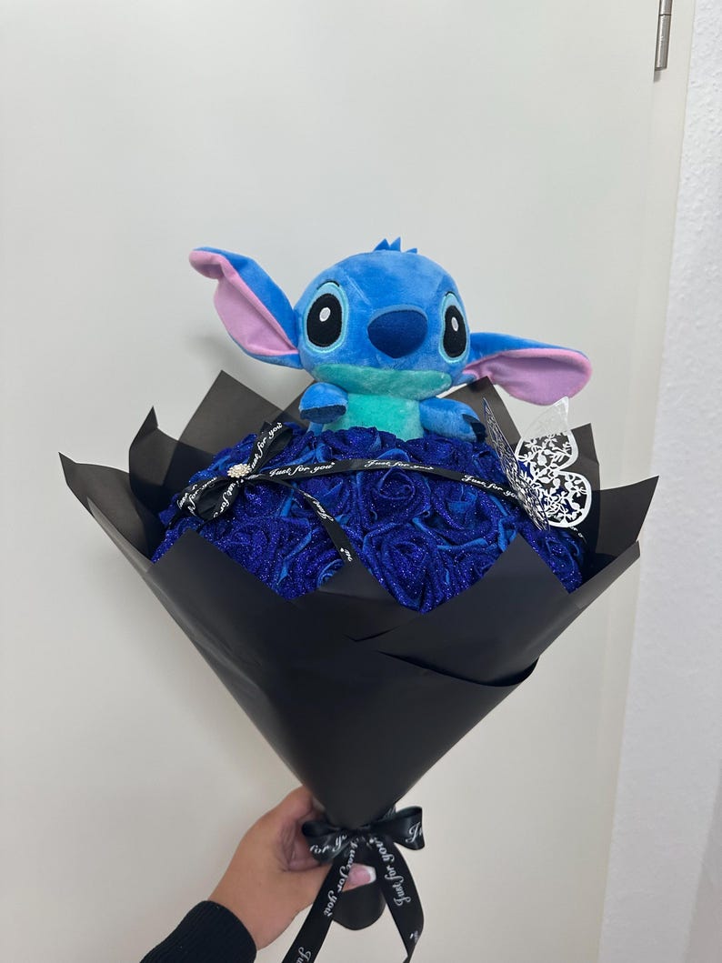 May include: A bouquet featuring a blue plush Stitch toy atop a bed of blue roses, wrapped in black paper. The bouquet is tied with a black ribbon that says "Just for you". A silver heart-shaped ornament is attached.