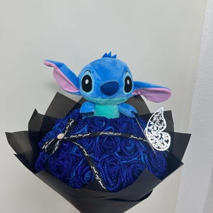 May include: A bouquet featuring a blue Stitch plush toy atop a bed of blue glitter roses. The arrangement is wrapped in black paper and tied with a ribbon that reads "Just for you." A decorative silver butterfly adds a delicate touch.