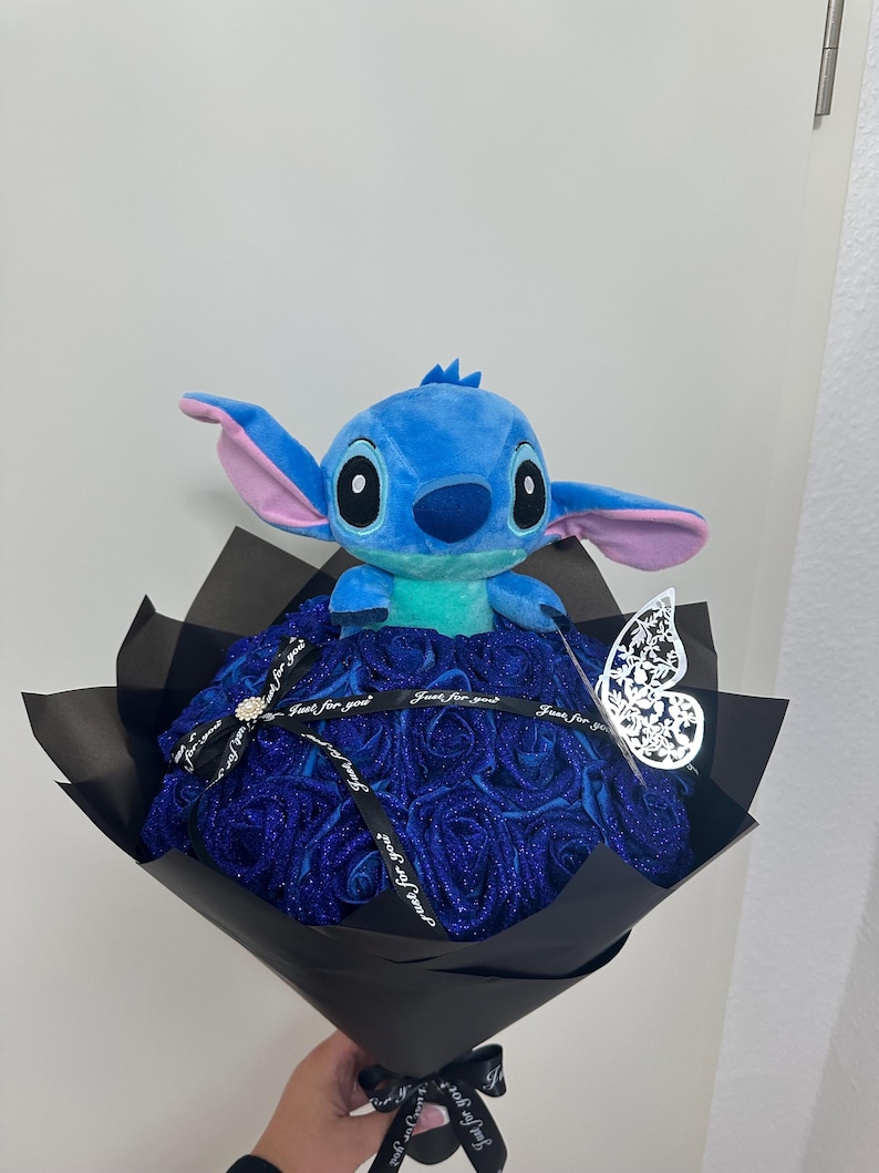 May include: A bouquet featuring a blue Stitch plush toy atop a bed of blue glitter roses. The arrangement is wrapped in black paper and tied with a ribbon that reads "Just for you." A decorative silver butterfly adds a delicate touch.