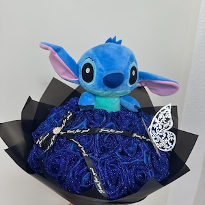 May include: A plush blue Stitch character sits atop a bouquet of blue glitter roses. The bouquet is wrapped in black paper and tied with a ribbon that says "Just for you". A decorative butterfly is attached.