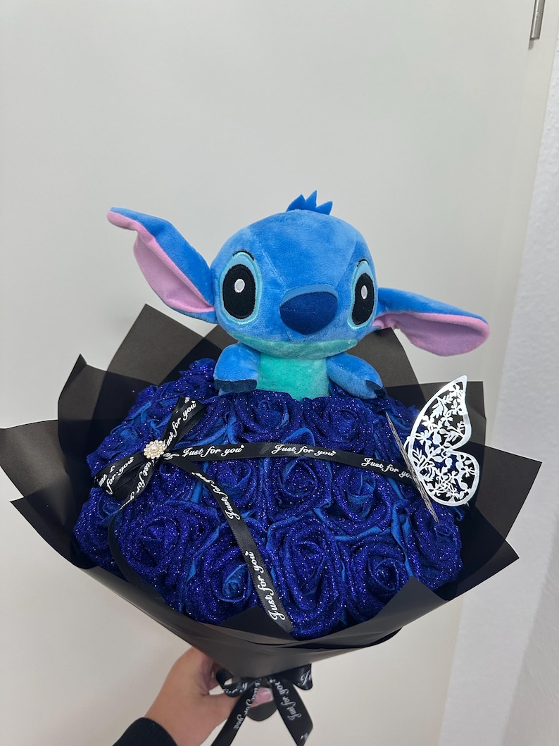 May include: A plush blue Stitch character sits atop a bouquet of blue glitter roses. The bouquet is wrapped in black paper and tied with a ribbon that says "Just for you". A decorative butterfly is attached.