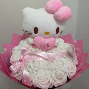 Op de afbeelding: Een boeket met een witte Hello Kitty-knuffel die een roze hart met de tekst "Love" vasthoudt. Het boeket bestaat uit witte rozen en is verpakt in roze papier met een roze lint.