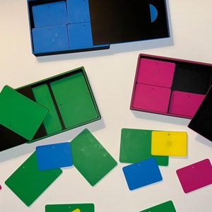 Può includere: Assortimento di piastrelle quadrate colorate con un piccolo foro al centro. Le piastrelle sono organizzate in tre scatole aperte con coperchi neri. Le scatole sono in plastica e hanno un colore diverso per ogni set di piastrelle: blu, verde e rosa.