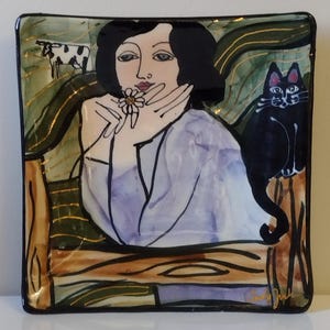 Może przedstawiać: Kwadratowy ceramiczny talerz z ręcznie malowanym wzorem. Grafika przedstawia kobietę trzymającą kwiat, czarnego kota i krowę. Talerz ma czarną obwódkę i błyszczące wykończenie. Paleta kolorów obejmuje odcienie niebieskiego, zielonego i brązowego.