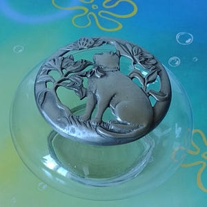 Puede incluir: Un cuenco de vidrio transparente con una figura decorativa de gato de metal plateado en la parte superior. El gato está sentado y rodeado de hojas y flores. El fondo es un degradado de azul y verde.