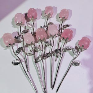 Pode incluir: Uma coleção de grampos de cabelo em forma de rosa. Cada grampo apresenta uma cabeça de quartzo rosa, haste prateada e detalhes de folhas. Os grampos estão dispostos sobre uma superfície branca com um fundo roxo suave.