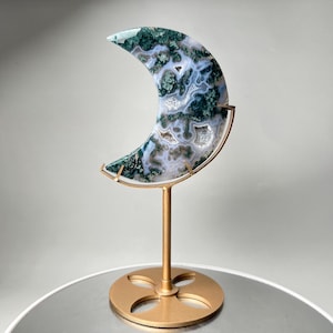 May include: A crescent moon-shaped moss agate stone featuring intricate green and white patterns with small crystalline inclusions, mounted on a gold-colored metal stand with a circular base and a four-petal cutout design.