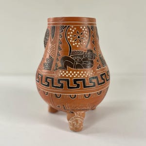 May include: A terracotta-colored, three-legged vase with intricate black and white geometric and figurative designs. The vase features a rounded body and a slightly flared rim. The base of each leg is a carved face.