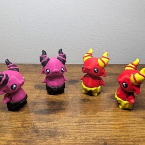 May include: Four small, 3D printed figurines of cartoon-like creatures with horns and tails. Two are pink with black horns and tails, and two are red with yellow horns and tails.