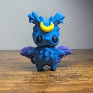 Może przedstawiać: Niebieska figurka z żółtym półksiężycem na głowie i fioletowymi skrzydłami. Figurka ma duże czarne oczy i szczegółowy, teksturowany wzór. Stoi na drewnianej powierzchni.