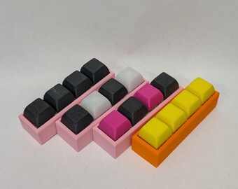 May include: A set of keyboard keycaps in various colors, including black, white, pink, and yellow. The keycaps are arranged in a staggered pattern and are housed in pink and orange plastic cases.