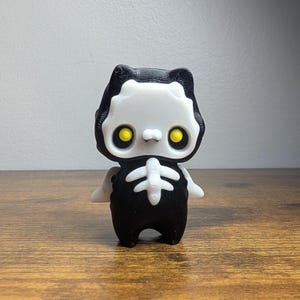 Puede incluir: Una pequeña figura de un gato esqueleto impresa en 3D, de color negro y blanco, con ojos amarillos. El gato lleva un disfraz negro con un diseño de esqueleto blanco.