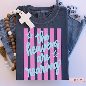 May include: A navy blue t-shirt with a pink striped design and the words "the heavens are roaring" in light blue script. The shirt is folded next to a pair of blue jeans and a wooden cross.