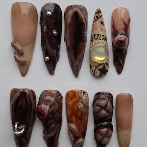 Könnte beinhalten: Zehn künstliche Fingernägel mit unterschiedlichen Designs. Die Nägel zeigen Brauntöne, Beige und Dunkelrot, mit Verzierungen wie silbernen Nieten, einem Stern- und Mondmuster und einem strukturierten Holzmaserungseffekt. Anordnung in einer Reihe.