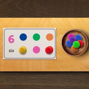 May include: Wooden educational toy featuring a card with the number 6 and the word "six" in pink. Six colored dots and six fuzzy balls in a wooden bowl are also present. The colors include blue, green, orange, yellow, pink, and red.