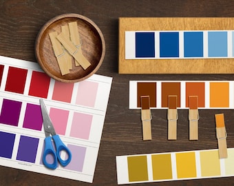 May include: An overhead shot of color swatches and wooden clothespins on a dark wood surface. The swatches display shades of red, purple, blue, brown, orange, and yellow. A pair of blue-handled scissors and a small wooden bowl are also present.