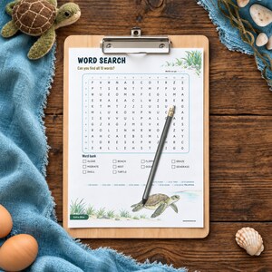 Puede incluir: Un portapapeles de madera con una hoja de sopa de letras titulada WORD SEARCH, un banco de palabras y una ilustraci&oacute;n de tortuga marina. Un l&aacute;piz gris descansa diagonalmente sobre la cuadr&iacute;cula. Una peque&ntilde;a tortuga de peluche y dos huevos marrones.