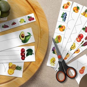 Pode incluir: Um conjunto de 50 tiras de papel com ilustrações coloridas de várias frutas, incluindo abacaxis, abacates, cerejas e bagas. As tiras têm linhas pontilhadas para cortar e traçar, acompanhadas por uma tesoura preta e laranja.