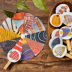 Könnte beinhalten: Ein farbenfroher, segmentierter Fächer und eine Holzschale mit Bastelstäbchen zum Thema Fische. Der Fächer weist verschiedene Muster und Farben auf, während die Stäbchen verschiedene Fischillustrationen zeigen. Eine lebendige, künstlerische Darstellung.