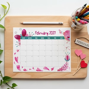 May include: A February 2027 calendar page featuring pink and purple watercolor illustrations of hearts, a butterfly, and a bird. The calendar includes a white pencil, a glass jar filled with colorful crayons, and three heart-shaped decorative picks.