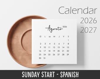 May include: A square white calendar page for August 2026 with black text, featuring the month name Agosto, the year 2026, and a grid of dates starting on Sunday. The calendar is partially overlapping a round, matte terracotta-colored ceramic saucer.